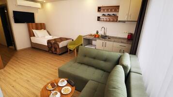 A hotel room with a bed, a green sofa, a small kitchen area, and a TV mounted on the wall.