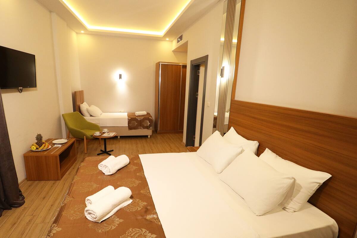 A hotel room with a large bed, a green armchair, a small table with a fruit plate, and a TV mounted on the wall.