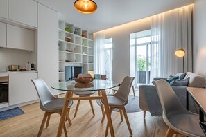 Et moderne køkken med hvide skabe, et spiseområde med et glasbord og grå stole, samt en opholdsstue med en sofa og et fjernsyn.