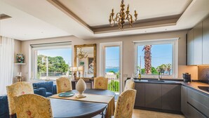Un coin repas avec une table ronde, des chaises à motifs floraux et un lustre. De grandes fenêtres offrent une vue sur la verdure et un bâtiment.