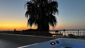 Une terrasse sur le toit offrant une vue sur un coucher de soleil au-dessus d’une étendue d’eau, avec un palmier et un espace piscine.