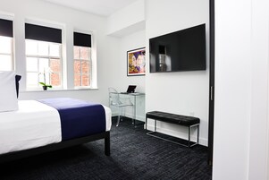 Una habitación de hotel moderna con una cama, un escritorio con una computadora portátil, un banco y un televisor en la pared.