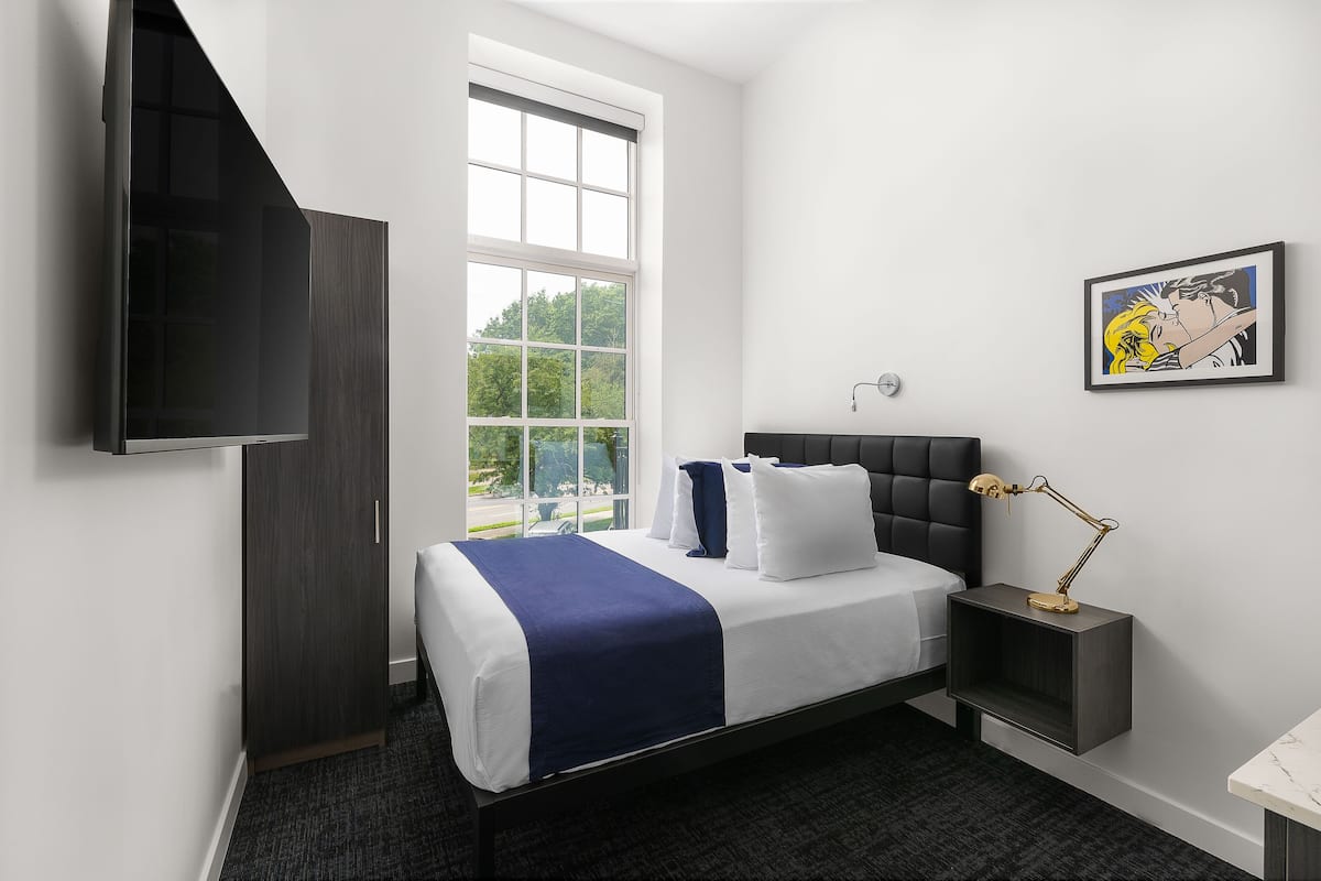 A modern hotel room with a large window, a flat-screen TV, a bedside table with a lamp, and a framed picture on the wall.