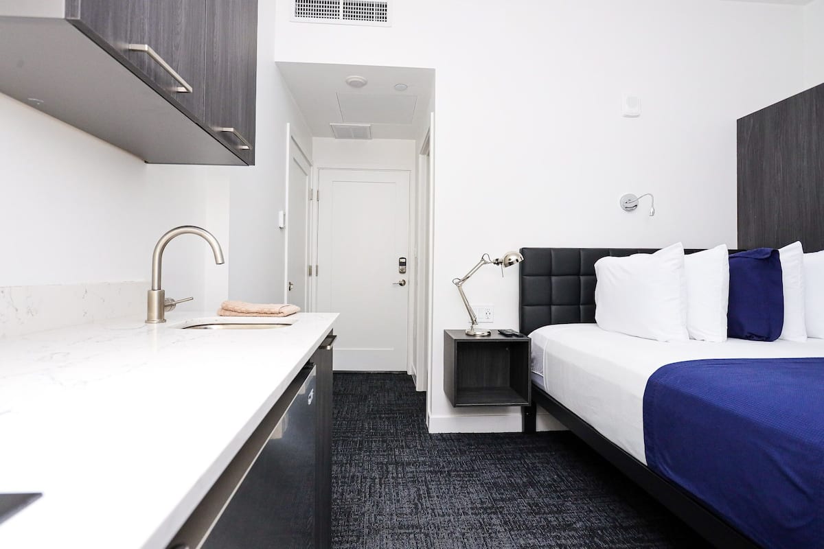 A modern hotel room with a white sink, a black upholstered bed, and a small bedside table with a lamp.