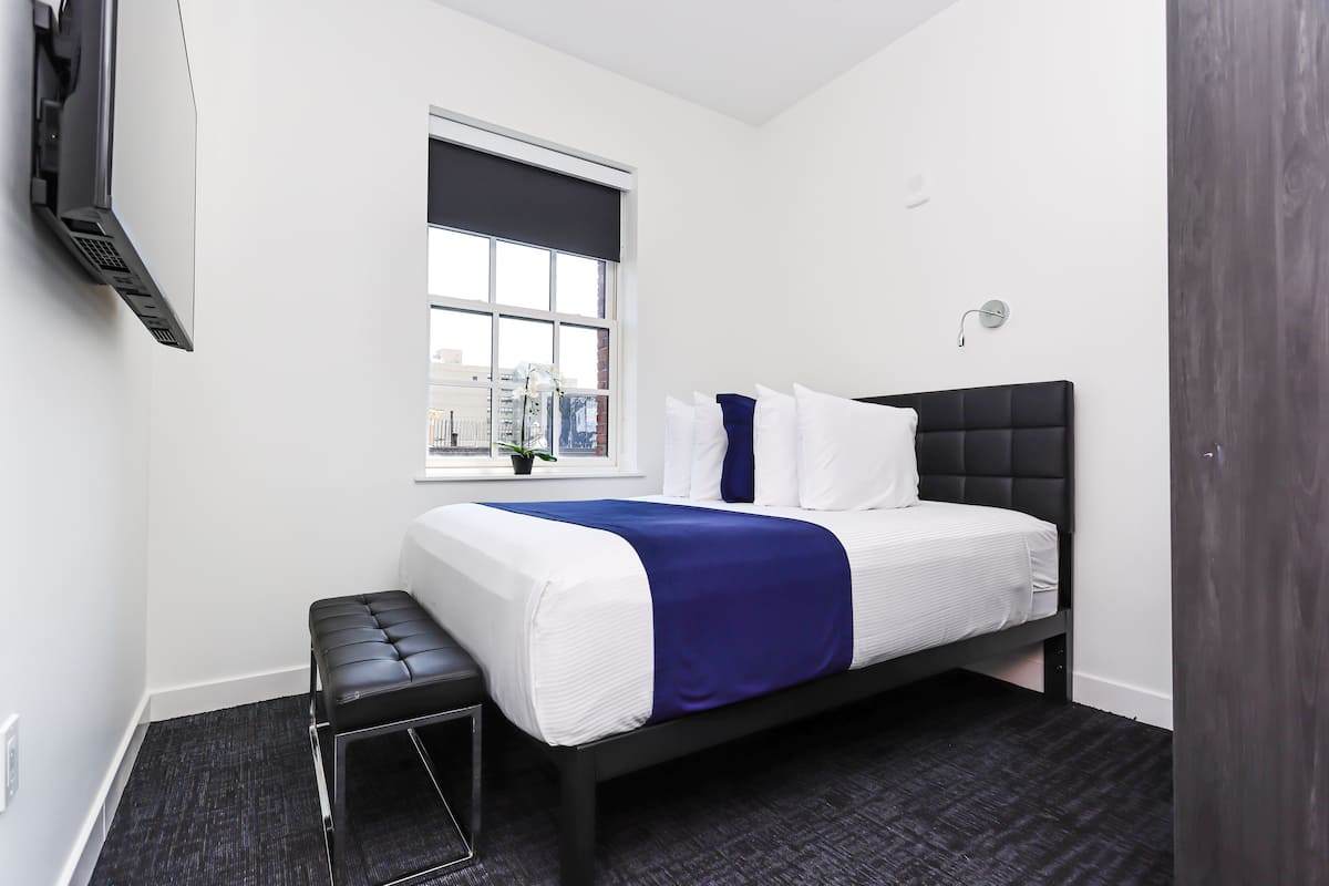 A modern hotel room with a bed, a bench, a window with a city view, and a mounted TV.