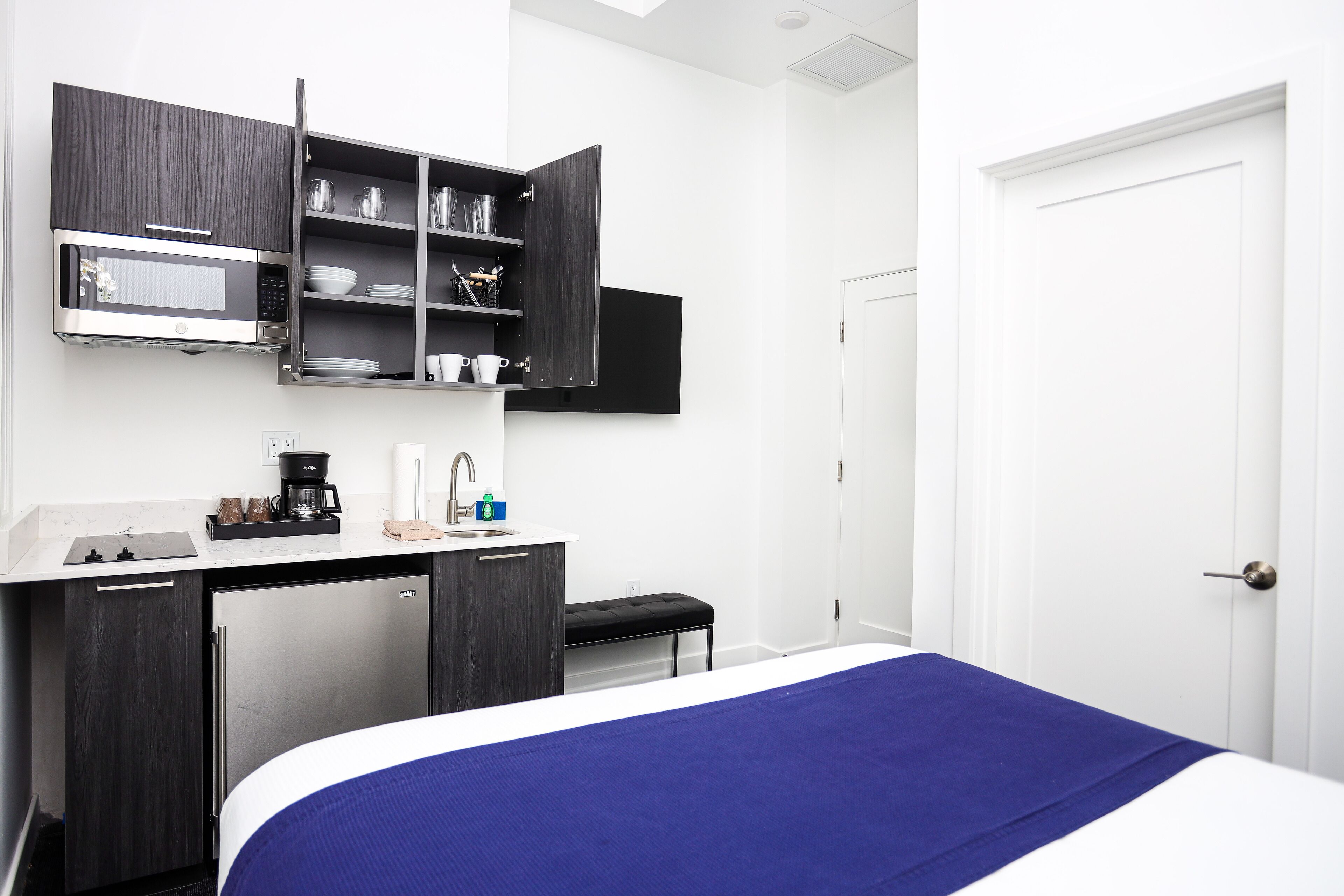 A compact hotel room with a bed, a desk with a microwave, a coffee maker, and a small refrigerator.