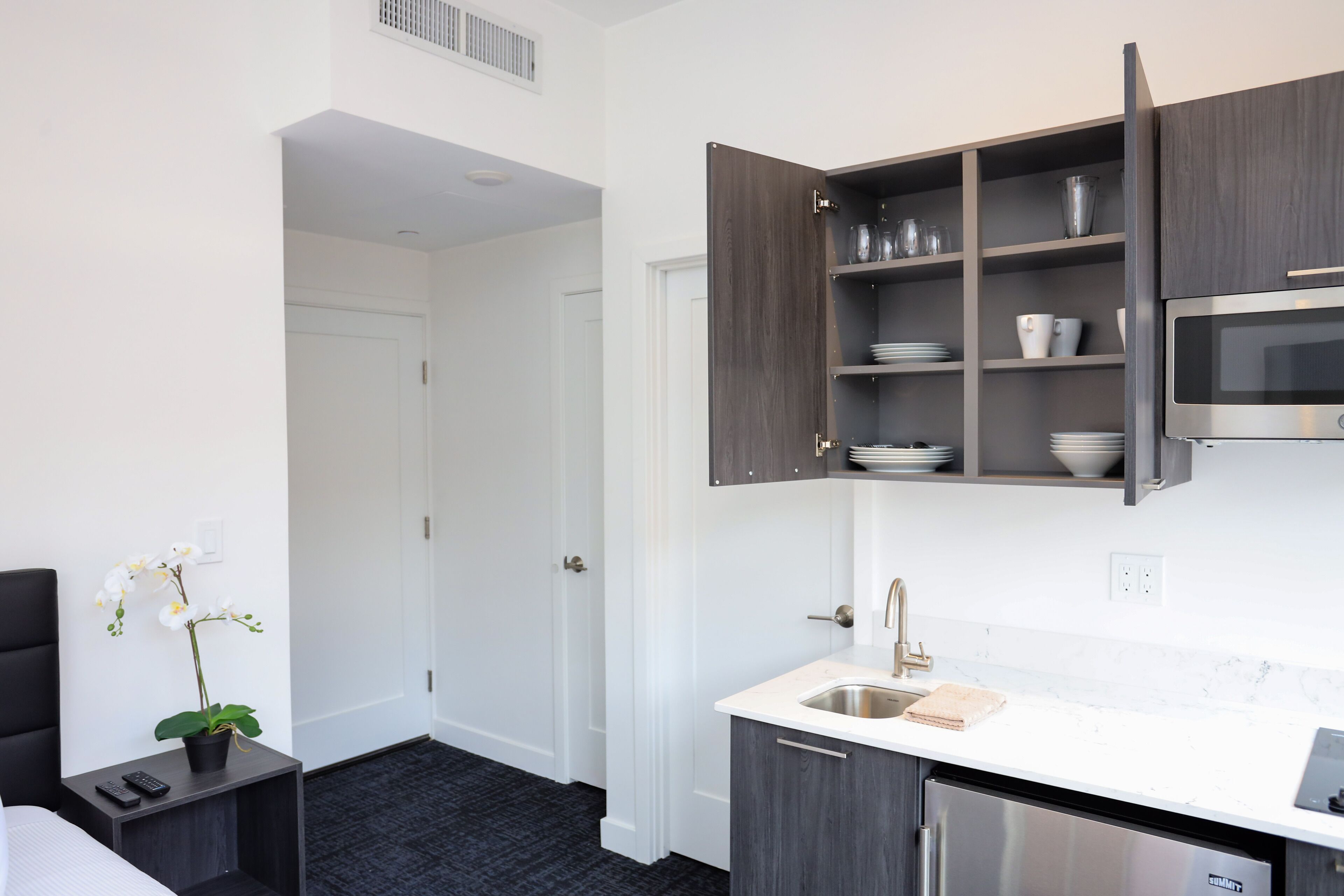 A compact hotel kitchen with a built-in microwave, a sink, and dark wooden cabinets.