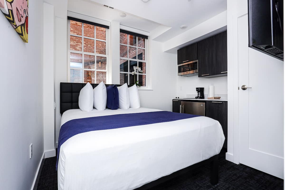 A hotel room with a large bed, a small kitchenette, and a window with a view of brick buildings.