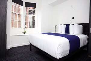 A modern hotel room with a large bed, a window with a view of a brick building, a small potted plant, and a wall-mounted light fixture.