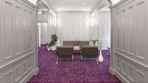 Un couloir aux murs recouverts de panneaux blancs, un tapis violet, deux chaises, un vase blanc et une plante.