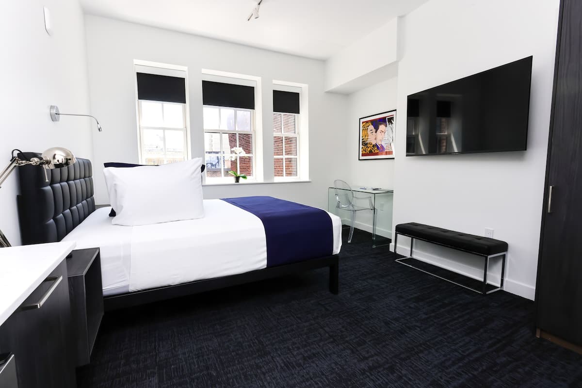 A modern hotel room with a bed, bedside table, desk, chair, TV, and windows.