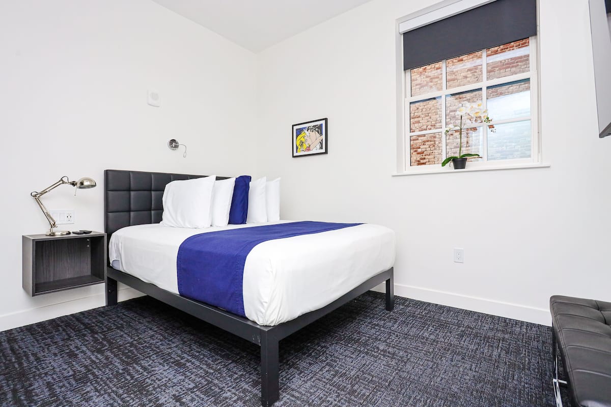 A modern bedroom with a bed, bedside table, lamp, and a window with a view of a brick wall.