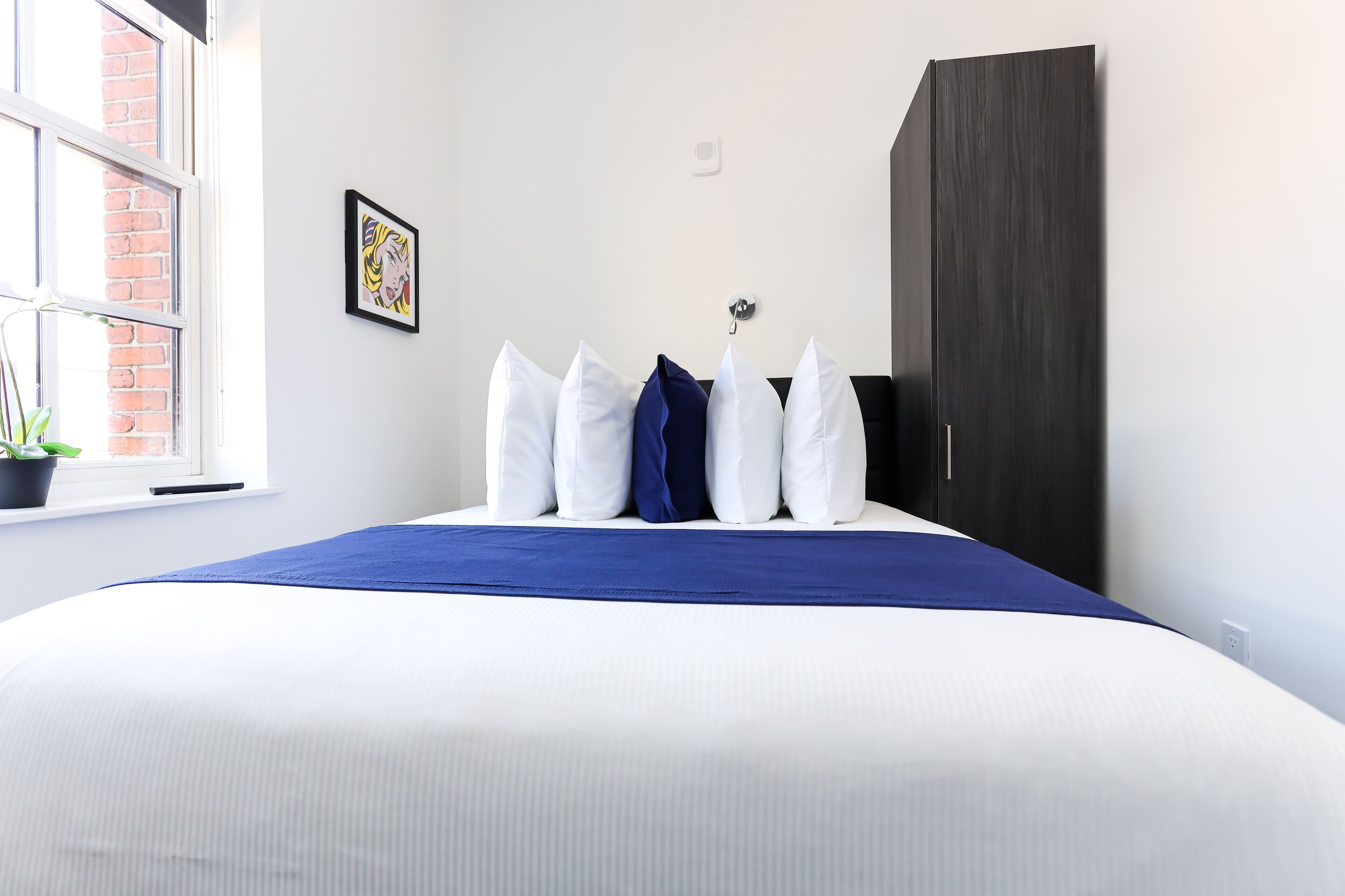 Cama bem arrumada com roupa de cama branca e azul, um guarda-roupa escuro, um quadro na parede e uma janela com uma planta.