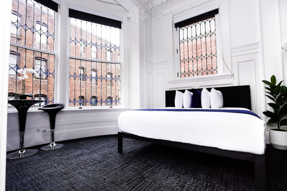 A modern bedroom with a large bed, two black bar stools, a potted plant, and a view of a brick building through a window with decorative iron bars.