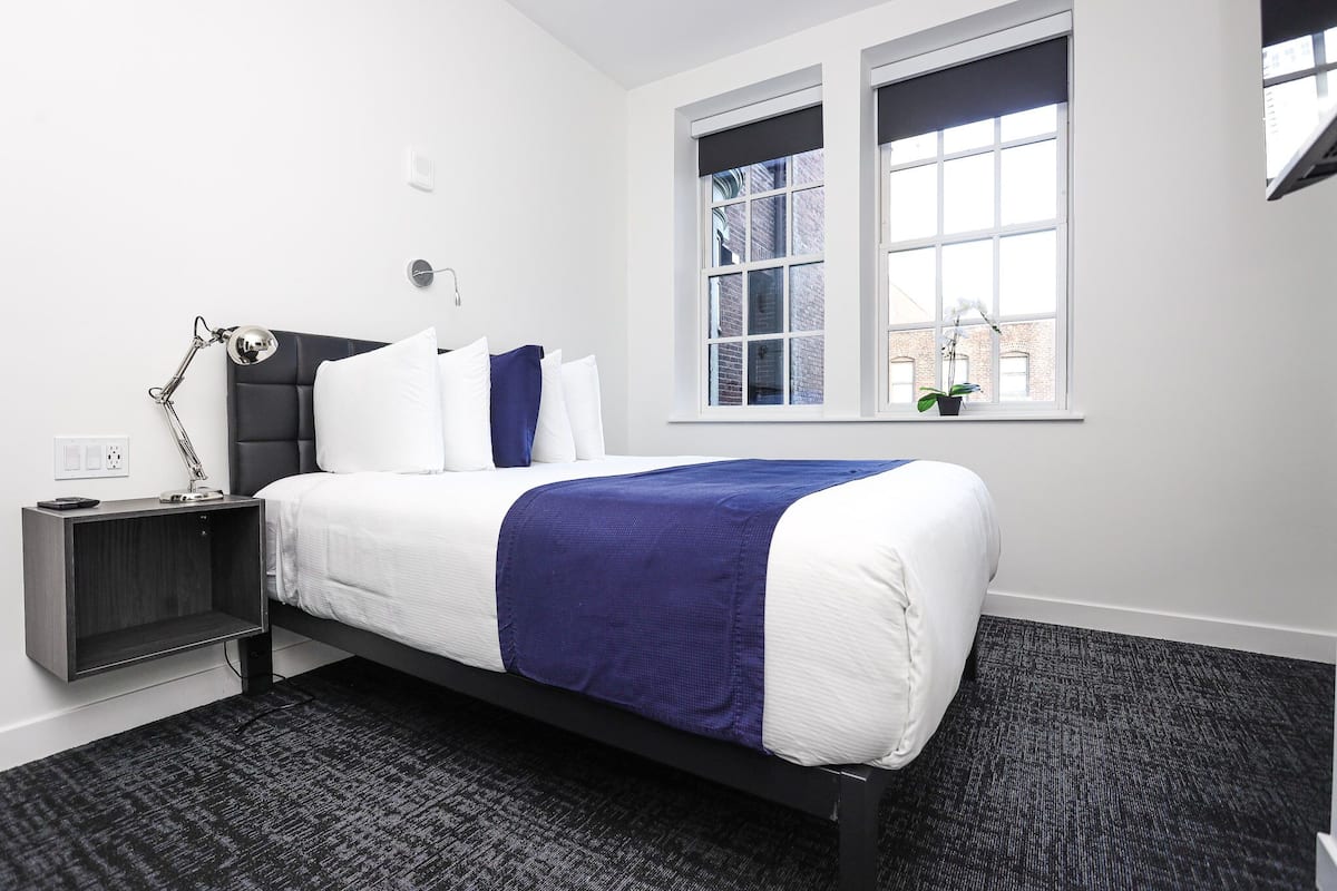 A modern bedroom with a bed, a nightstand, a lamp, and a window with a view of buildings.