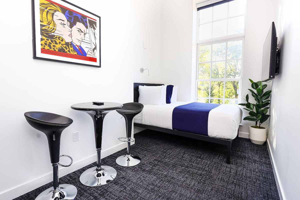 A hotel room with a bed, two bar stools, a small table, a TV mounted on the wall, and a window with a view of trees.
