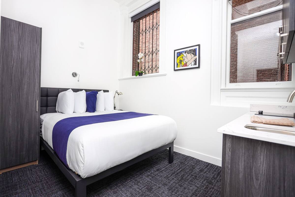 A bedroom with a bed, a window with a view of a brick building, a small plant on the windowsill, and a framed picture on the wall.