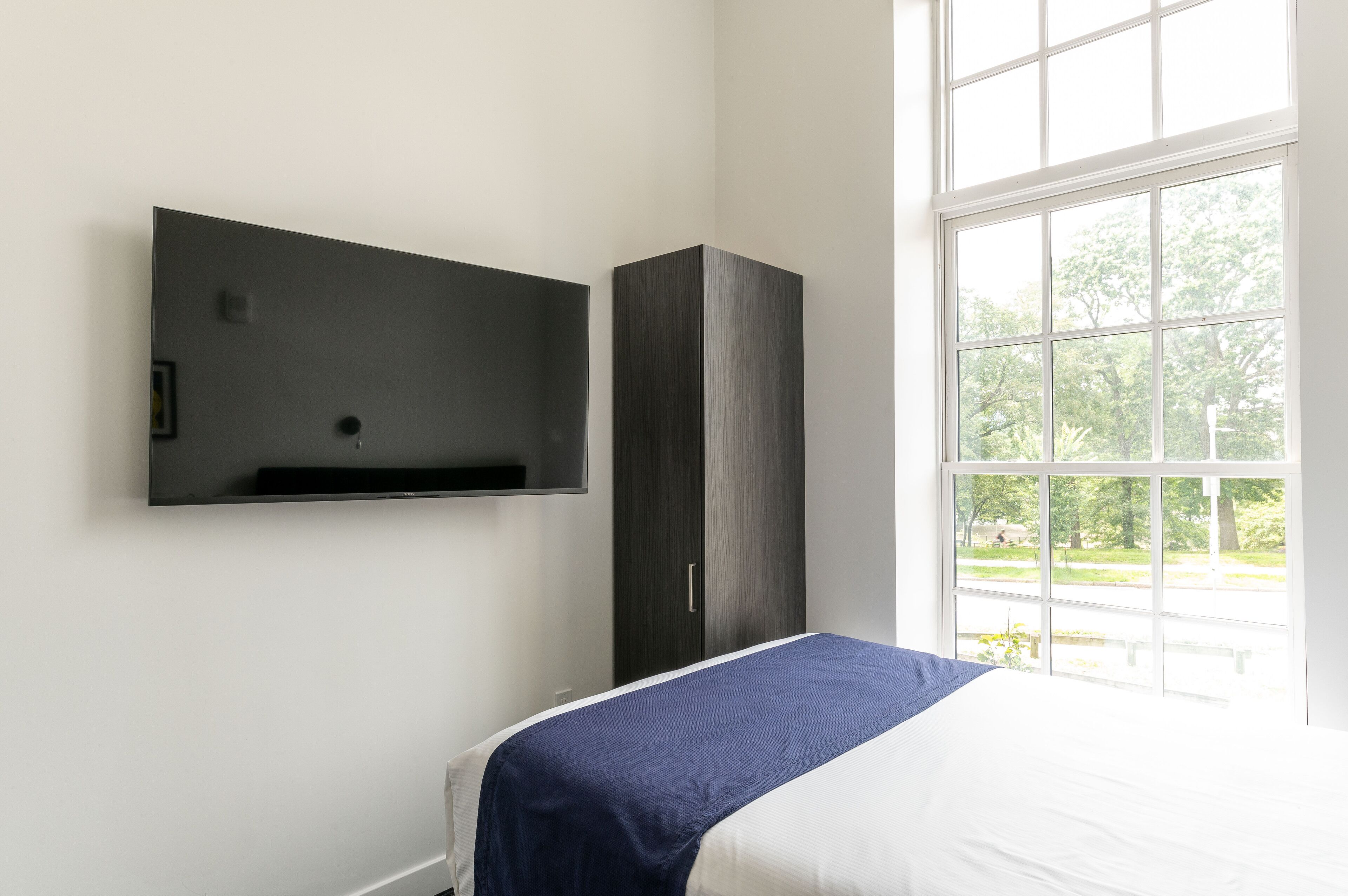 Une chambre à coucher comprenant un lit, une armoire, un téléviseur fixé au mur et une fenêtre donnant sur un paysage verdoyant.