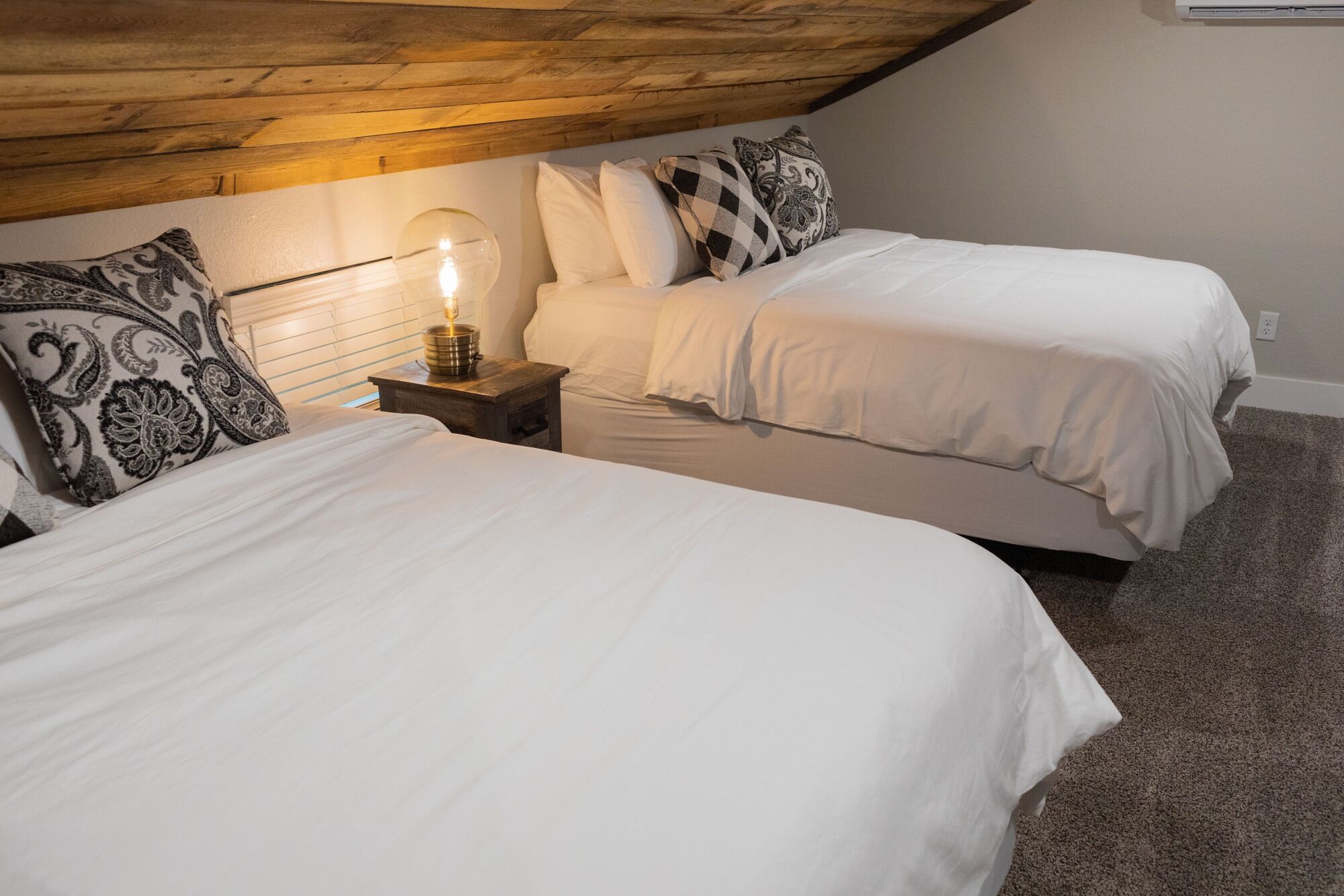 Un dormitorio con dos camas, una mesita de noche con lámpara y techo de madera.