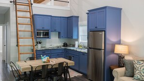 A compact kitchen with blue cabinets, a stainless steel refrigerator, a microwave, and a dining area with a wooden table and chairs. - Cozy, Spacious, and Right on the Banks of Rocky Creek, Two Creeks Crossing Resort (Livingston)