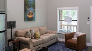 A living room with a beige sofa, a brown armchair, a dining table, and a painting of a boat on the wall. - Cozy, Spacious, and Right on the Banks of Rocky Creek, Two Creeks Crossing Resort (Livingston)