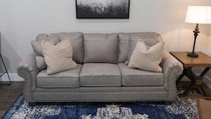 A grey sofa with two beige cushions, a blue patterned rug, a small table with a lamp, and a framed picture on the wall. - A Cozy Getaway Right on the Water, Two Creeks Crossing Resort (Livingston)