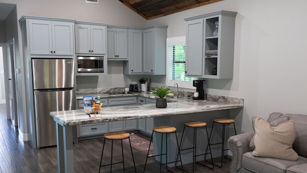 A modern kitchen with grey cabinets, stainless steel appliances, and a marble countertop. There are three bar stools and a grey sofa in the adjacent living area. - A Cozy Getaway Right on the Water, Two Creeks Crossing Resort (Livingston)