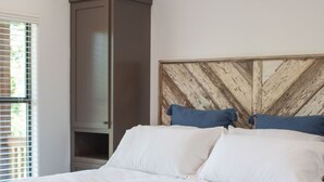 A neatly made bed with white linens, a wooden headboard, and a bench at the foot of the bed. - Cozy, Spacious, and Right on the Banks of Rocky Creek, Two Creeks Crossing Resort (Livingston)