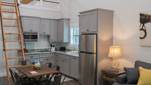Une cuisine compacte équipée d’un réfrigérateur, d’un four à micro-ondes et de placards. On y trouve une table à manger avec des chaises, ainsi qu’un petit canapé avec une lampe.
