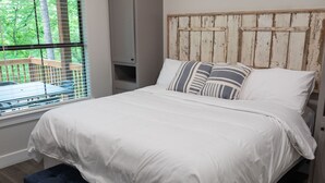 Une chambre avec un lit blanc, des coussins à rayures, une tête de lit en bois, un banc au pied du lit et une fenêtre avec des stores.
