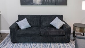Un sofá gris oscuro con dos almohadones estampados, una alfombra azul con diseño y una obra de arte de temática forestal en la pared.