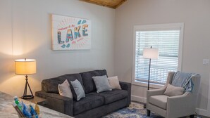 Una sala de estar con un sofá gris oscuro, un sillón gris claro, un tapete estampado azul y un cartel en la pared que dice "welcome to the LAKE stay awhile".