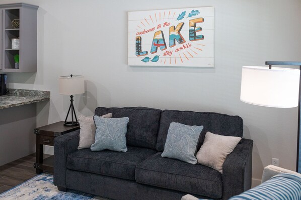 Un salón con un sofá gris oscuro, dos cojines estampados, una obra de arte en la pared blanca que dice "welcome to the LAKE stay awhile", una lámpara y una mesita auxiliar pequeña.