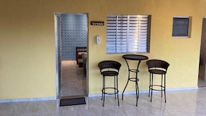 Un pasillo con una pared amarilla, una puerta que da a una habitación, un ventanal con persianas y tres sillas de mimbre.