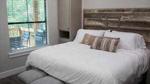Een slaapkamer met een bed, kussens, een bankje, een raam met jaloezieën en uitzicht op de omgeving.