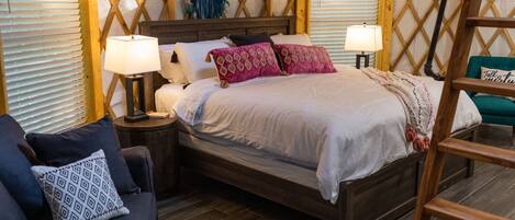 A cozy bedroom with a wooden bed, two bedside lamps, a woven wall hanging, and a blue patterned pillow on a dark sofa.