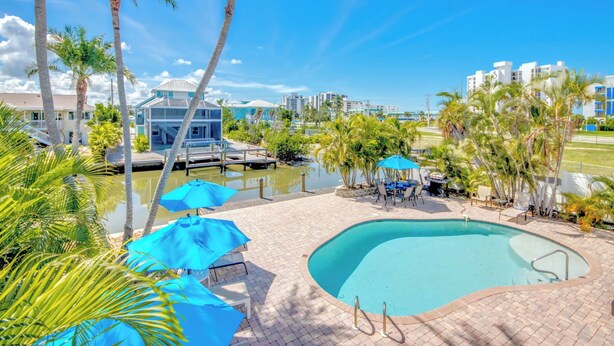 A swimming pool with a dock, palm trees, and a view of buildings in the background. - 110 Bahia #3 Pet Friendly Canal Unit with Pool (Fort Myers Beach)