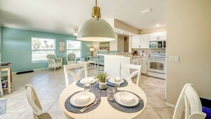 Un comedor con una mesa redonda dispuesta para dos, una cocina al fondo y una lámpara colgante grande sobre la mesa.