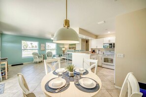 Un comedor con una mesa redonda dispuesta para dos, una cocina al fondo y una lámpara colgante grande sobre la mesa.