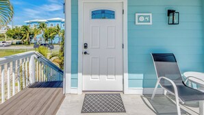 Une porte blanche avec une petite fenêtre, un balcon avec une chaise, et une vue sur un immeuble bleu bordé de palmiers.