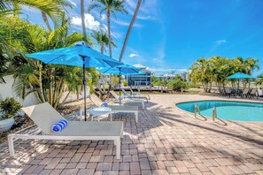 A poolside area with lounge chairs and umbrellas, palm trees, and a clear blue sky.