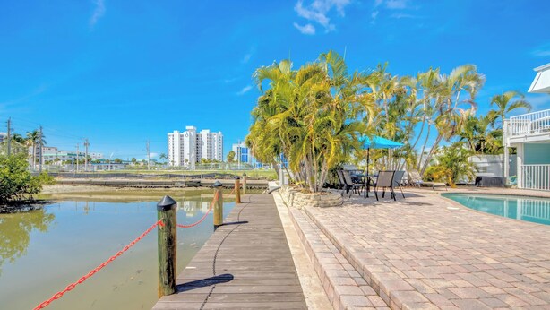 Un muelle de madera que da acceso a un área de piscina, con vistas a un cuerpo de agua tranquilo y un paisaje urbano al fondo.