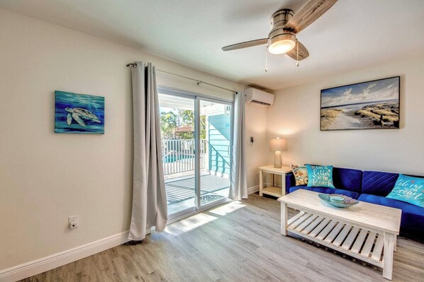 Una habitación con ventilador de techo, un cuadro con escena de playa, un sofá azul con almohadas de temática marina y una mesa de centro de madera.