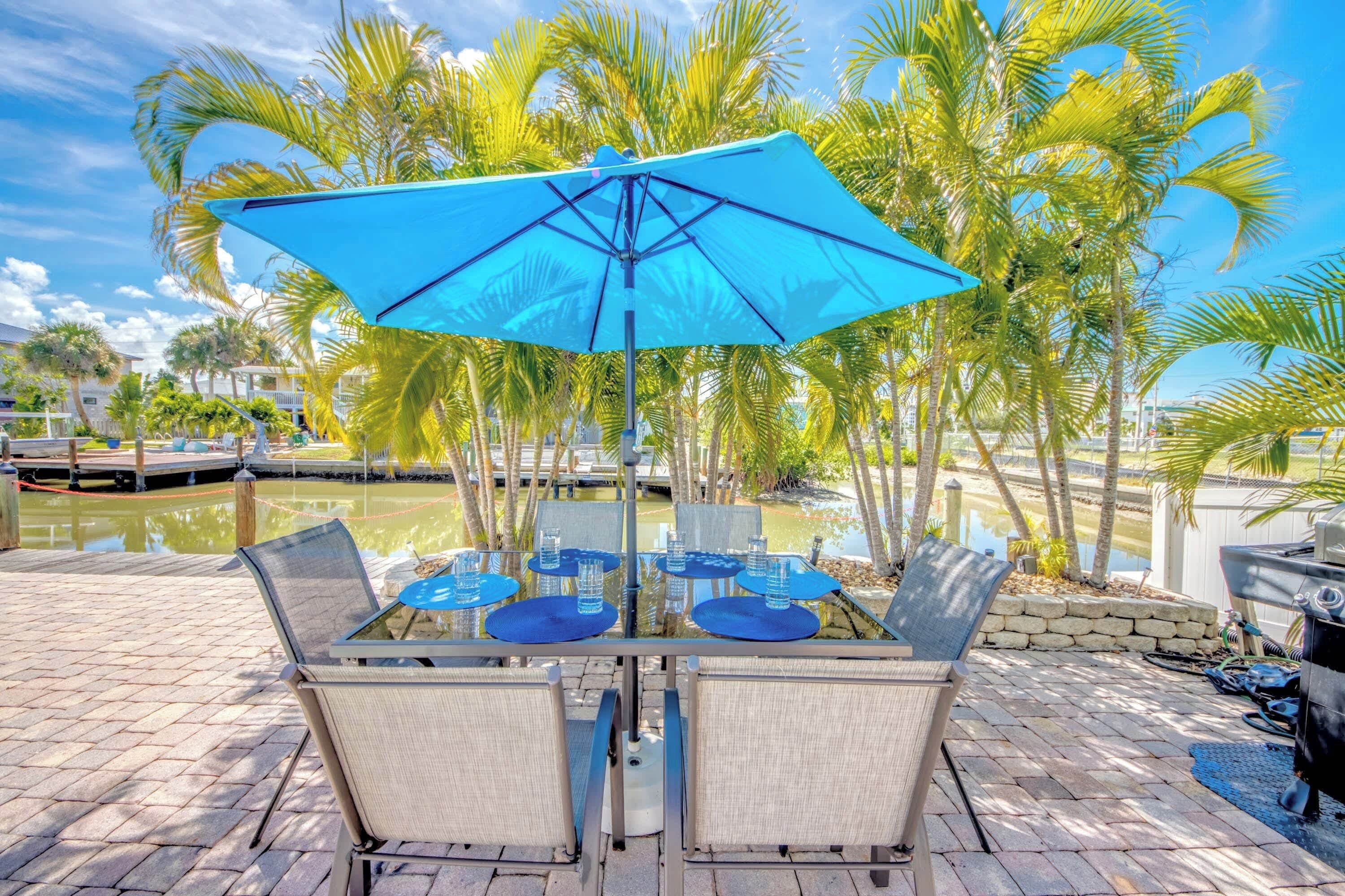 An outdoor dining area with a table and chairs, surrounded by palm trees and a body of water.