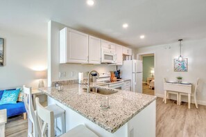 Una cocina moderna con armarios blancos, encimera de granito, electrodomésticos de acero inoxidable y una zona de comedor con mesa y sillas blancas.