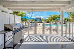 A covered outdoor area with a grill, a pool, and a view of a building with blue umbrellas.