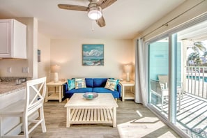 A room with a ceiling fan, a painting of a beach scene, a blue sofa with cushions, a coffee table, and a view of a pool through large windows.