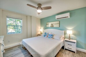 A bedroom with a bed, two bedside tables with lamps, a ceiling fan, an air conditioning unit, and a window with a view of palm trees.