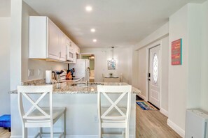 Una cocina moderna con armarios blancos, encimera de granito y una isla central con taburetes de bar. Hay una zona de comedor con mesa y sillas, y un elemento decorativo en la pared.