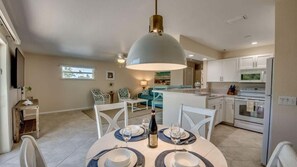 Un coin repas avec une table ronde dressée pour quatre personnes, un grand luminaire suspendu et une cuisine en arrière-plan.